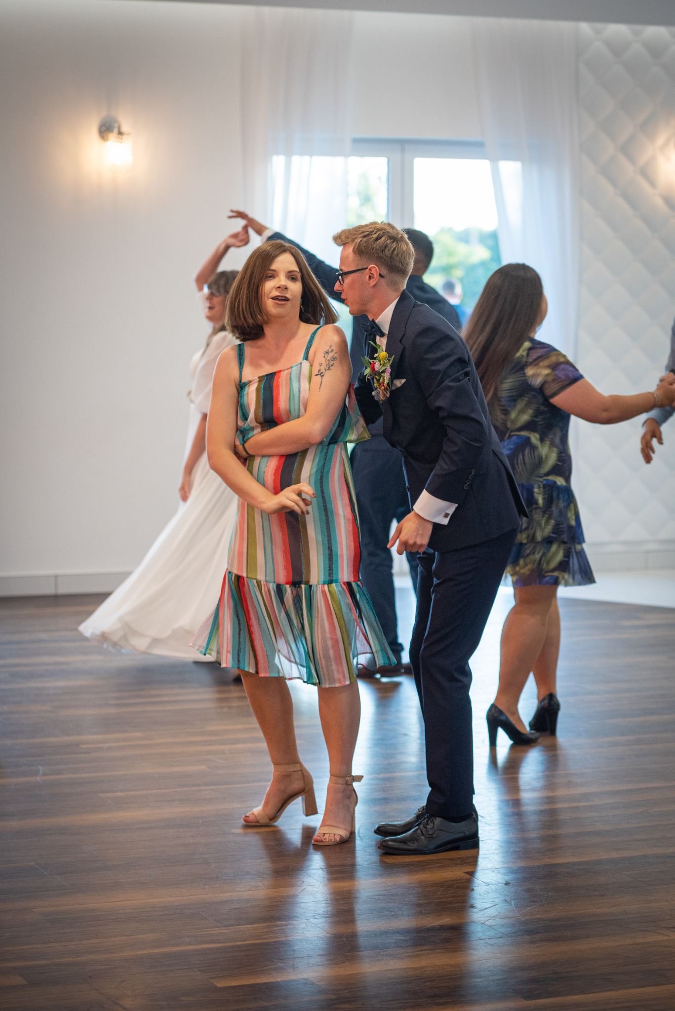 Goście weselni tańczą na parkiecie, kobieta w sukience w kolorowe paski i tatuażem na ramieniu, mężczyzna w granatowym garniturze z butonierką.