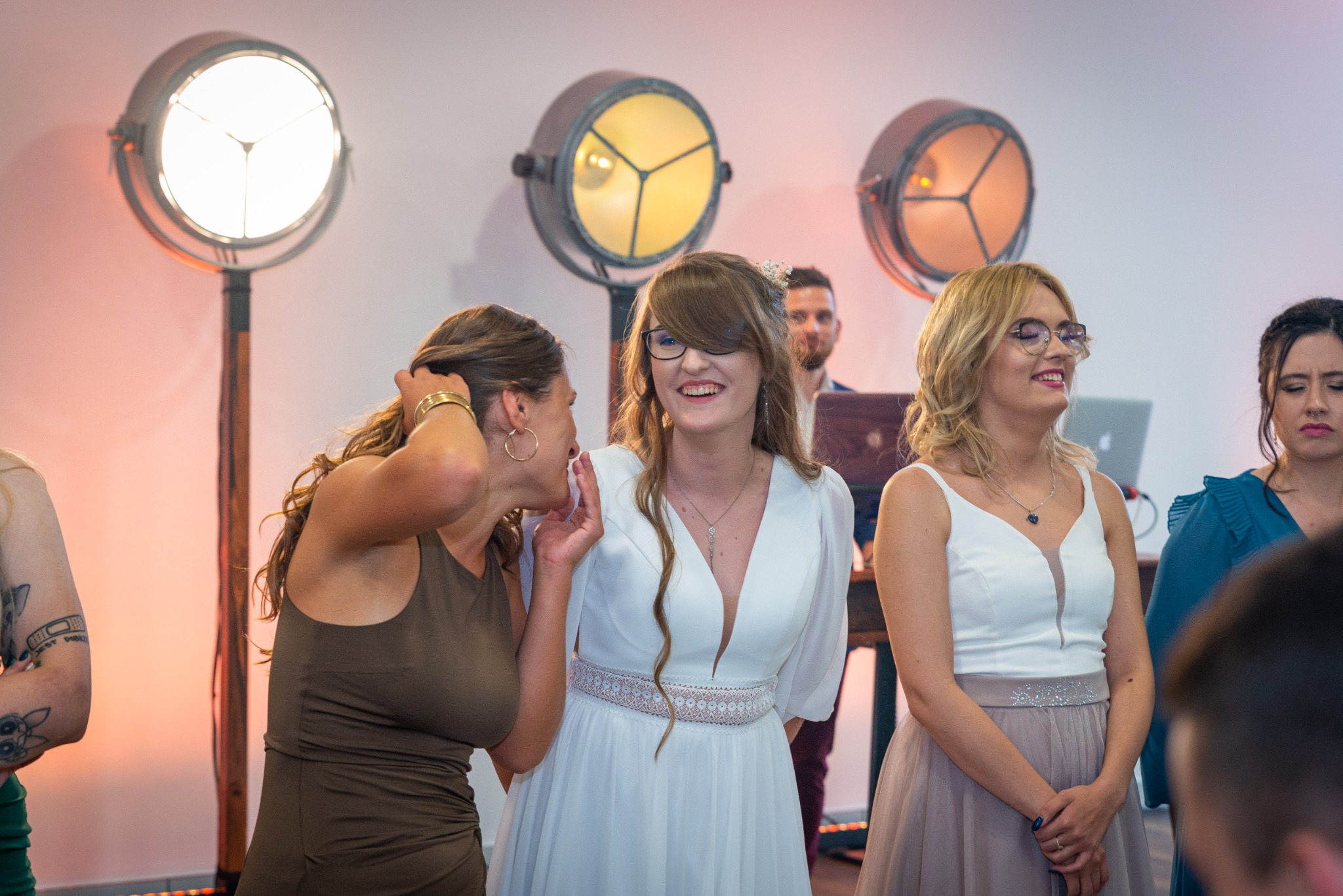 Uśmiechnięta panna młoda w białej sukni z koronkowym pasem, otoczona gośćmi weselnymi podczas przyjęcia, w tle oświetlenie sceniczne.