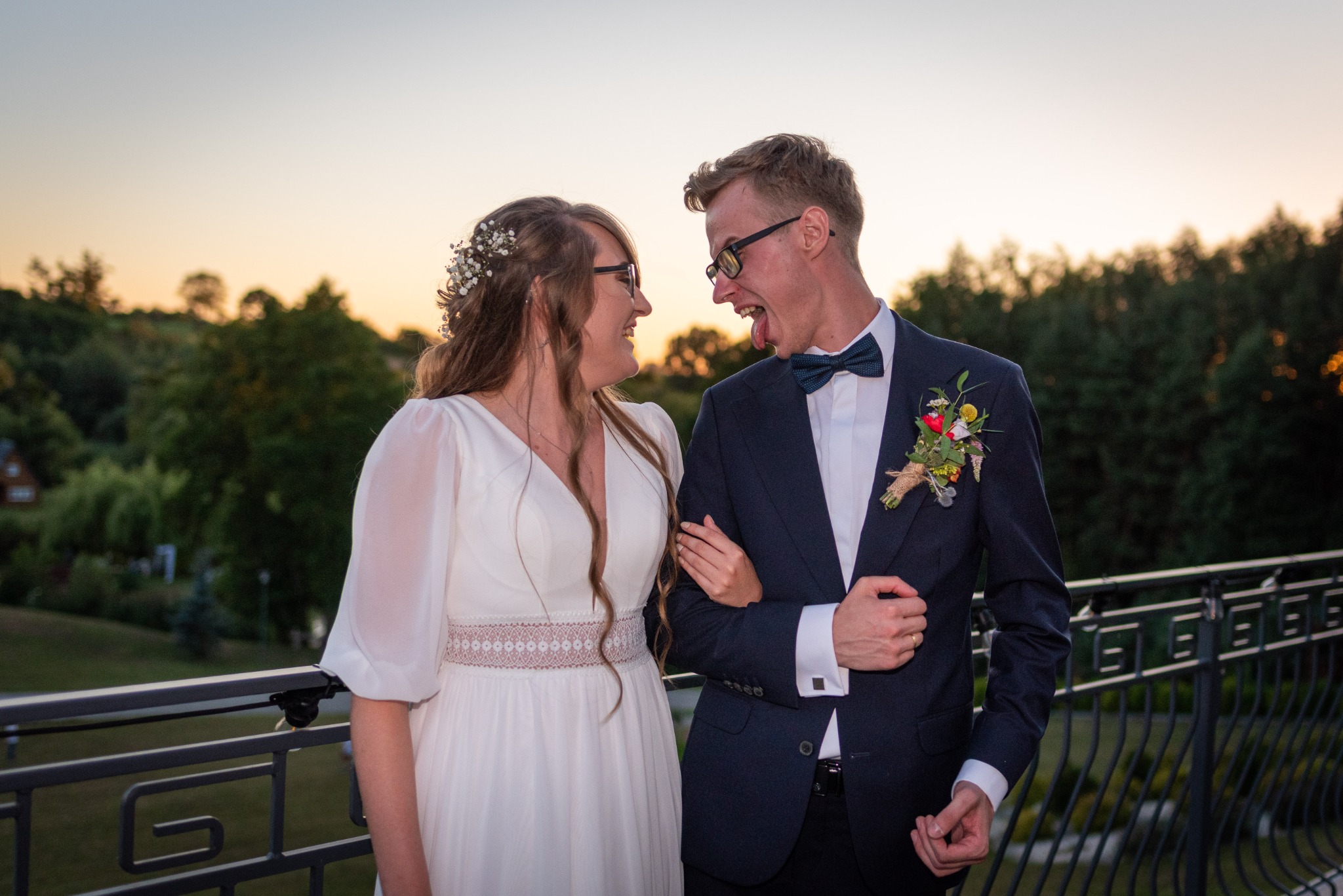 Para młoda na balkonie wpatruje się w siebie, pan młody w czarnych okularach pokazuje język, w tle zachód słońca i zieleń drzew.