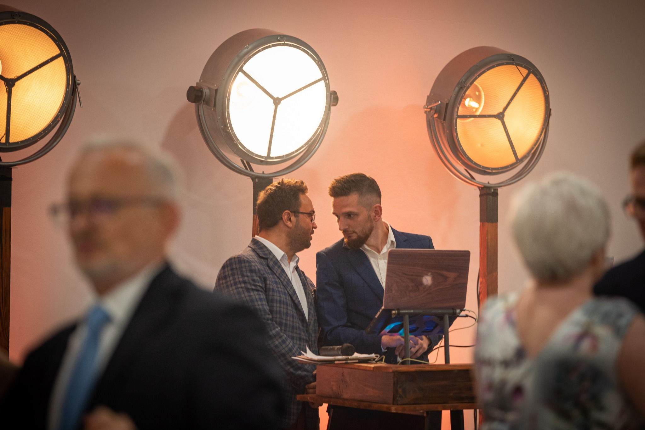 Dwaj mężczyźni w garniturach rozmawiają przy drewnianym pulpicie z laptopem, oświetleni przez industrialne lampy sceniczne na tle widowni.