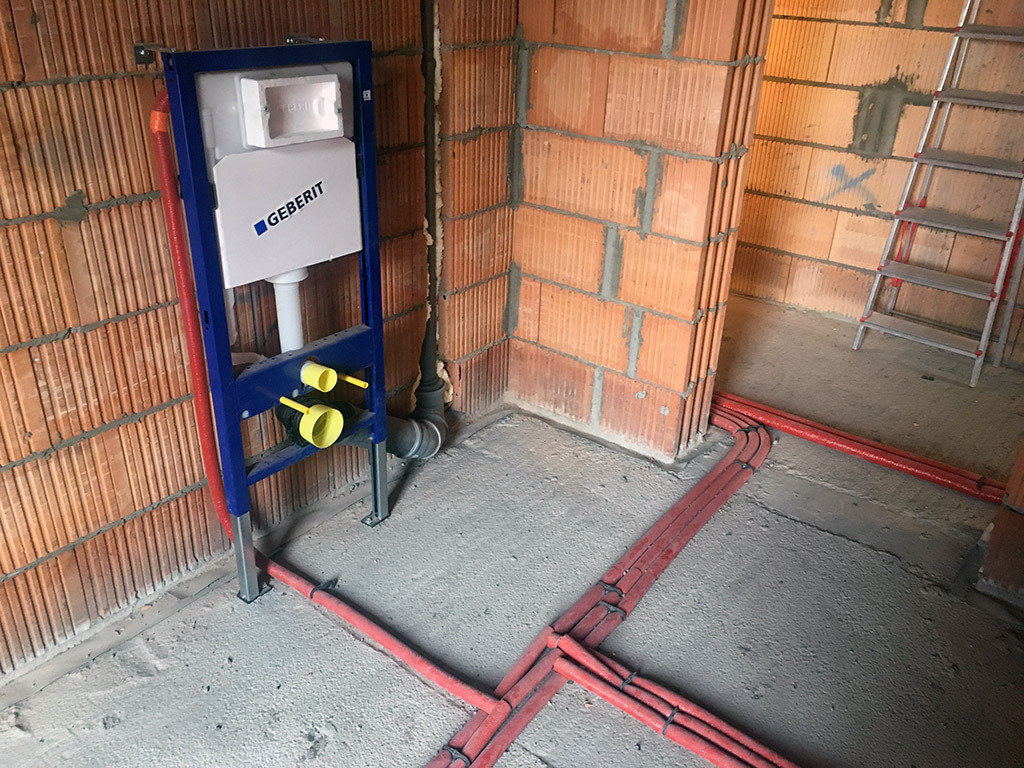 Stelaż podtynkowy Geberit zamontowany w surowym stanie, widoczne czerwone rury instalacji wodnej na posadzce, ściany z cegły dziurawki, drabina aluminiowa w tle.