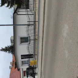 Biała elewacja domu jednorodzinnego z czarną, ozdobną metalową bramą i fragmentem chodnika oraz ulicy w kadrze.