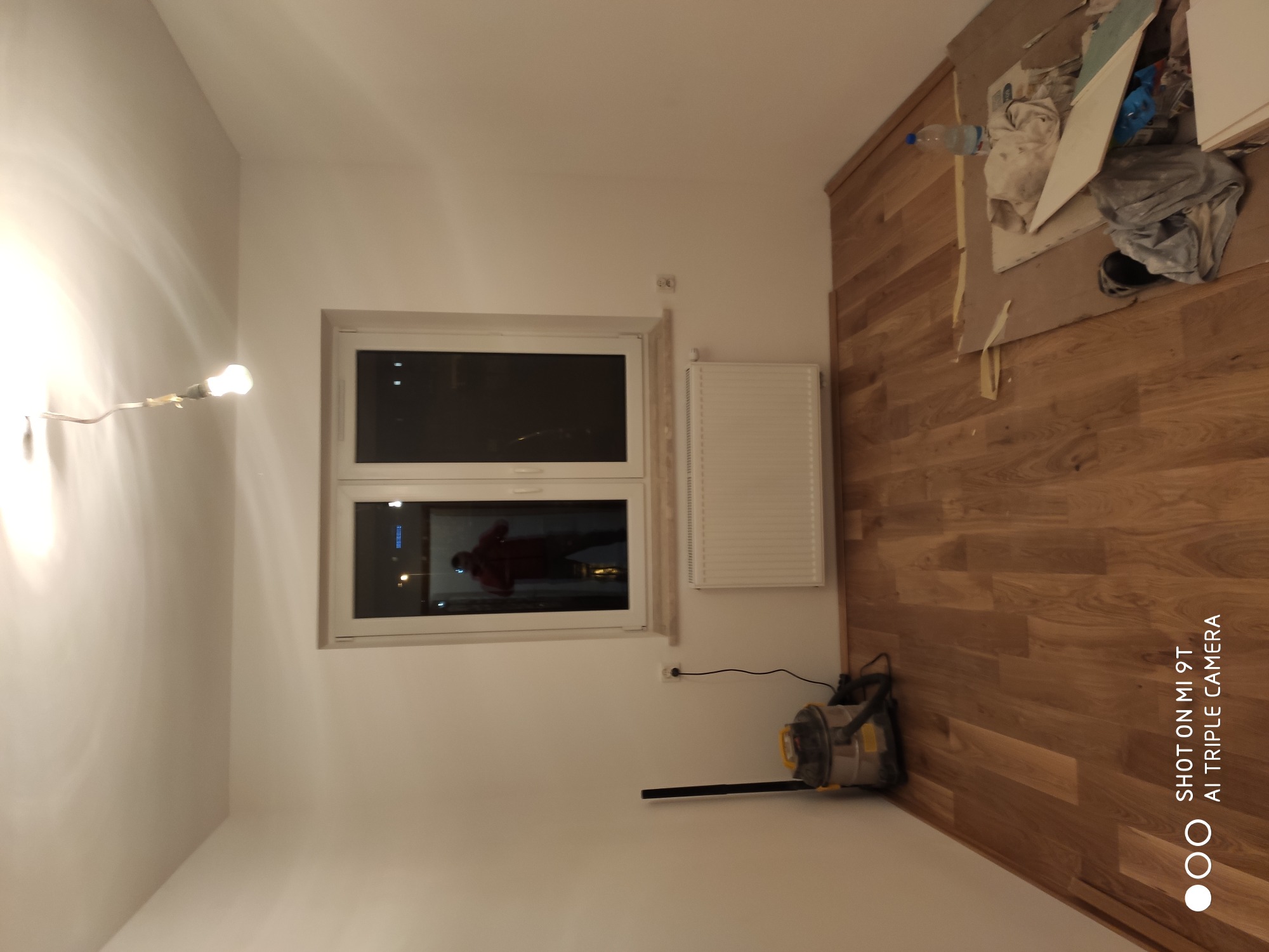 Pokój po remoncie: świeżo położona drewniana podłoga, pomalowane ściany na biało, widoczny grzejnik i okno z odbiciem osoby, odkurzacz przemysłowy w rogu pomieszczenia, widoczne pozostałości po...