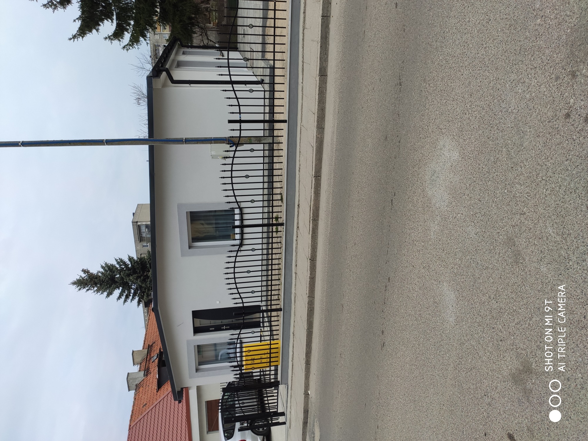 Biała elewacja domu jednorodzinnego z czarną, ozdobną metalową bramą i fragmentem chodnika oraz ulicy w kadrze.