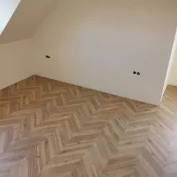 Jasne panele ułożone we wz&oacute;r jodełki w pokoju na poddaszu z białymi ścianami i widocznymi gniazdkami elektrycznymi.