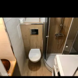 Nowoczesna łazienka z drewnopodobnymi płytkami, wiszącą toaletą z chromowanym przyciskiem spłukiwania, kabiną prysznicową z zaokrąglonymi drzwiami i pralką.