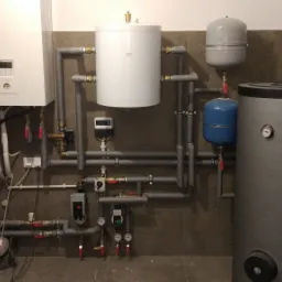Zestaw urządzeń grzewczych w kotłowni: wiszący kocioł gazowy, biały zasobnik ciepłej wody, szary zbiornik buforowy i niebieskie naczynia wzbiorcze, połączone szarymi rurami z zaworami i manometrami...