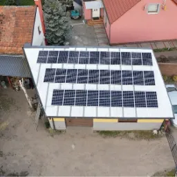 Widok z góry na instalację fotowoltaiczną składającą się z wielu paneli na dachu budynku gospodarczego, obok zaparkowany samochód osobowy.