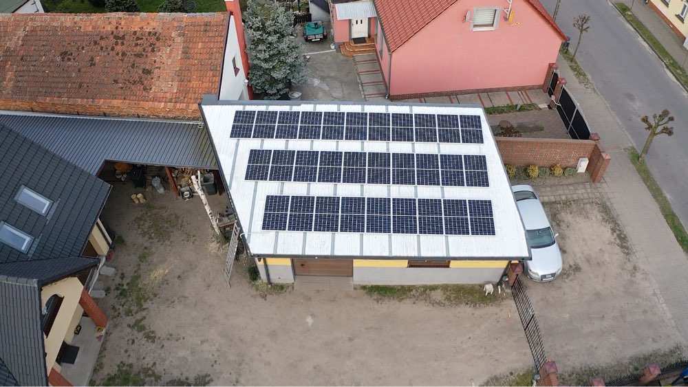 Widok z góry na instalację fotowoltaiczną składającą się z wielu paneli na dachu budynku gospodarczego, obok zaparkowany samochód osobowy.