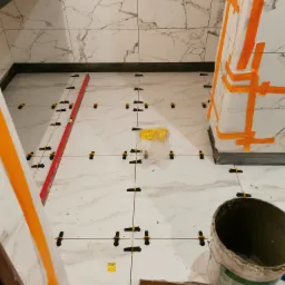 Białe płytki imitujące marmur ułożone na podłodze i ścianach łazienki, z użyciem systemu poziomowania płytek (klipsy i kliny), poziomica oraz obudowa pionu kanalizacyjnego zabezpieczona taśmą...