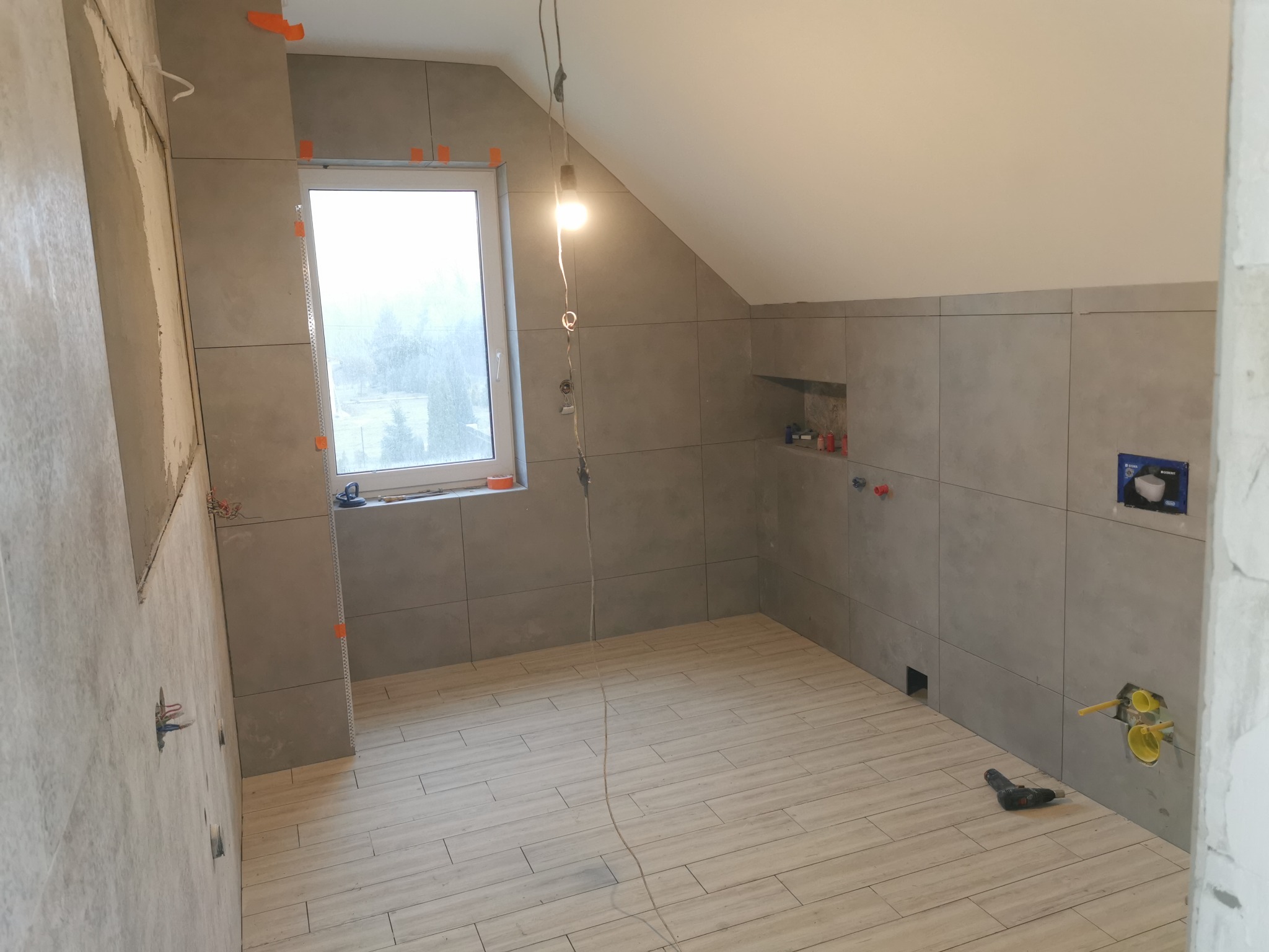 Łazienka w trakcie remontu na poddaszu: szare płytki na ścianach, drewnopodobna podłoga, widoczne instalacje wodne i elektryczne, okno z widokiem na drzewa, narzędzia na podłodze.