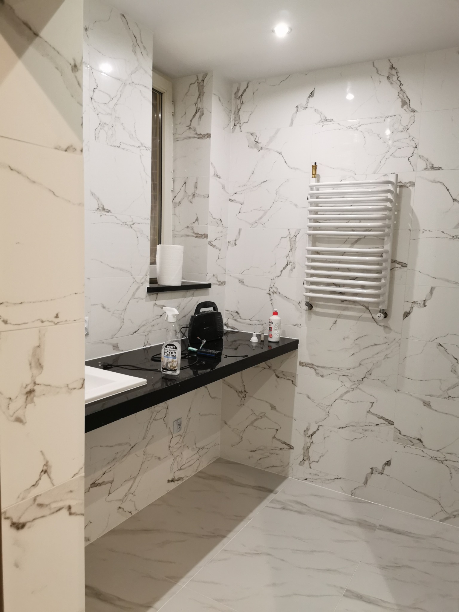 Biała łazienka wykończona płytkami imitującymi marmur, z czarnym blatem pod umywalką i grzejnikiem drabinkowym, widoczne akcesoria i środki czystości na blacie.