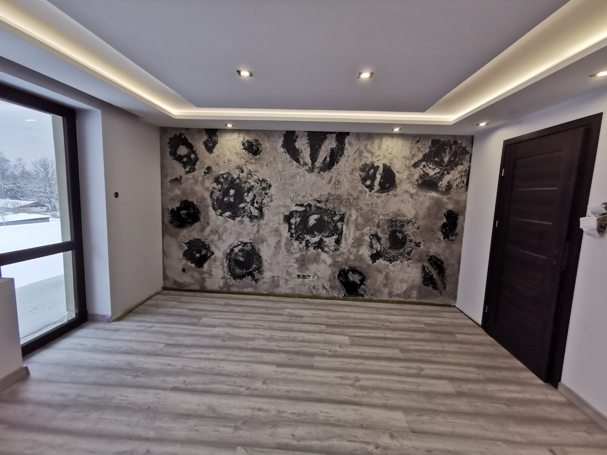 Pokój z oryginalną ścianą w stylu loft z szarym tłem i czarnymi, okrągłymi wzorami, panele podłogowe w odcieniach szarości, nowoczesne oświetlenie sufitowe LED i ciemne drzwi.