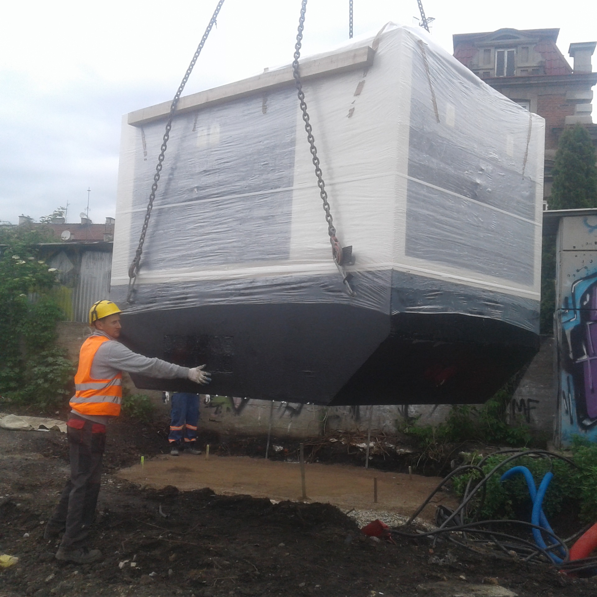 Pracownik w kasku i kamizelce odblaskowej asekuruje zawieszony na łańcuchach, owinięty folią modułowy budynek przenośny nad przygotowanym fundamentem z wystającymi prętami, w tle budynek mieszkalny...