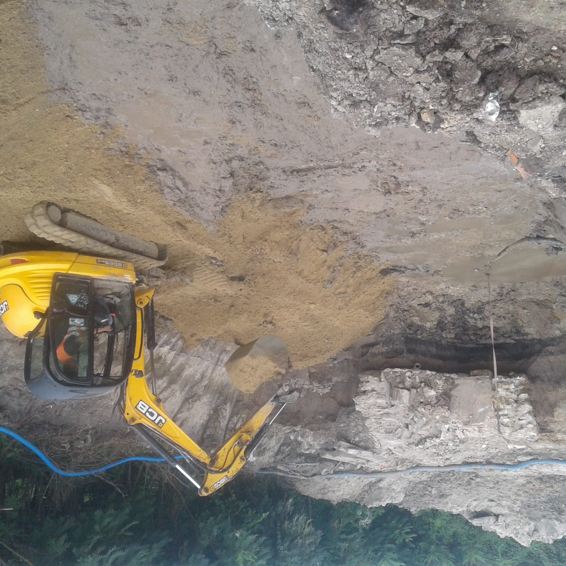 Żółta koparka JCB w trakcie wykopu ziemi w wykopie, widoczny fragment muru z kamienia i cegły oraz niebieski przewód.