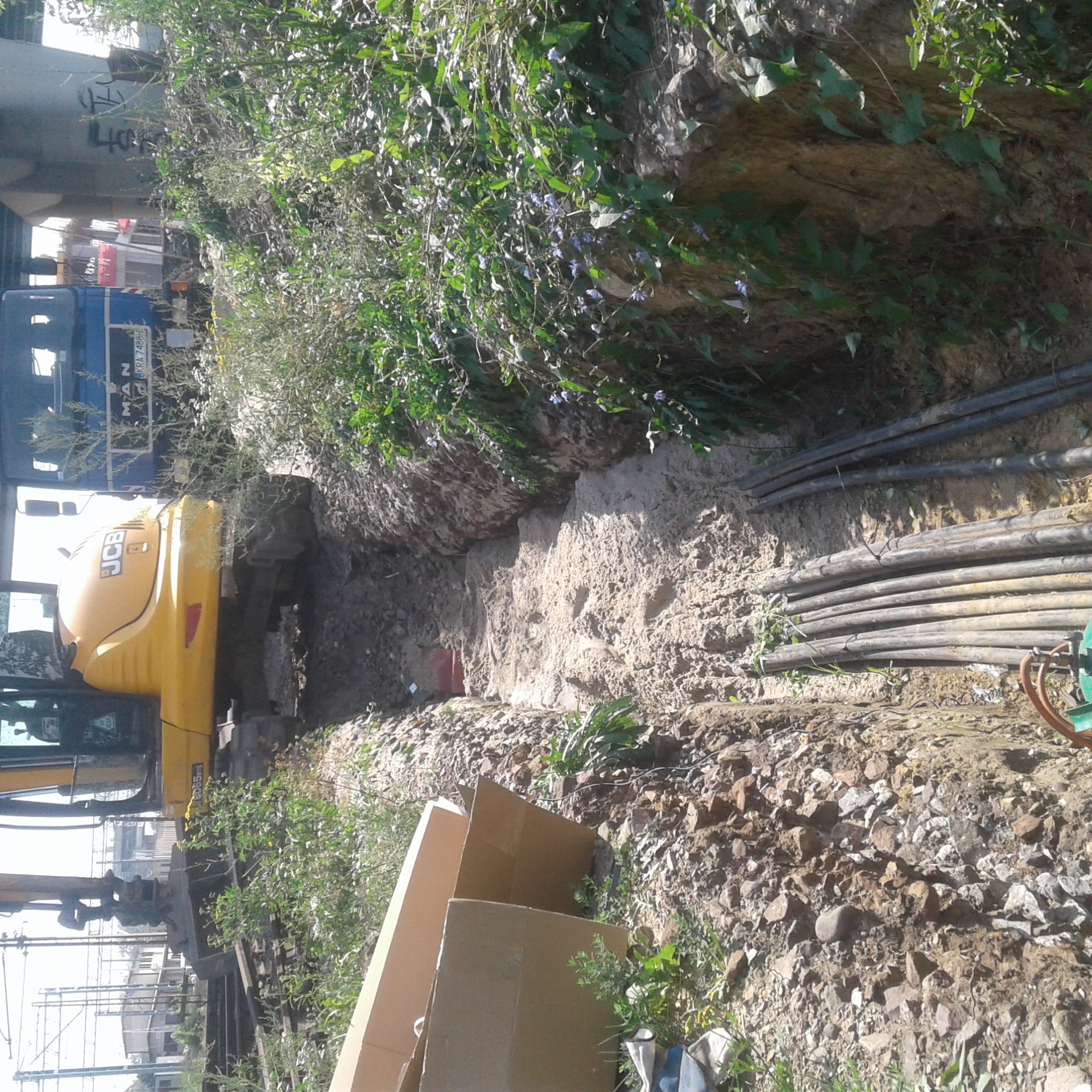 Żółta koparka JCB przy wykopie, w tle widoczny most i ciężarówka MAN, ułożone rury w wykopie, ziemia kamienista, zarośla, kartony.