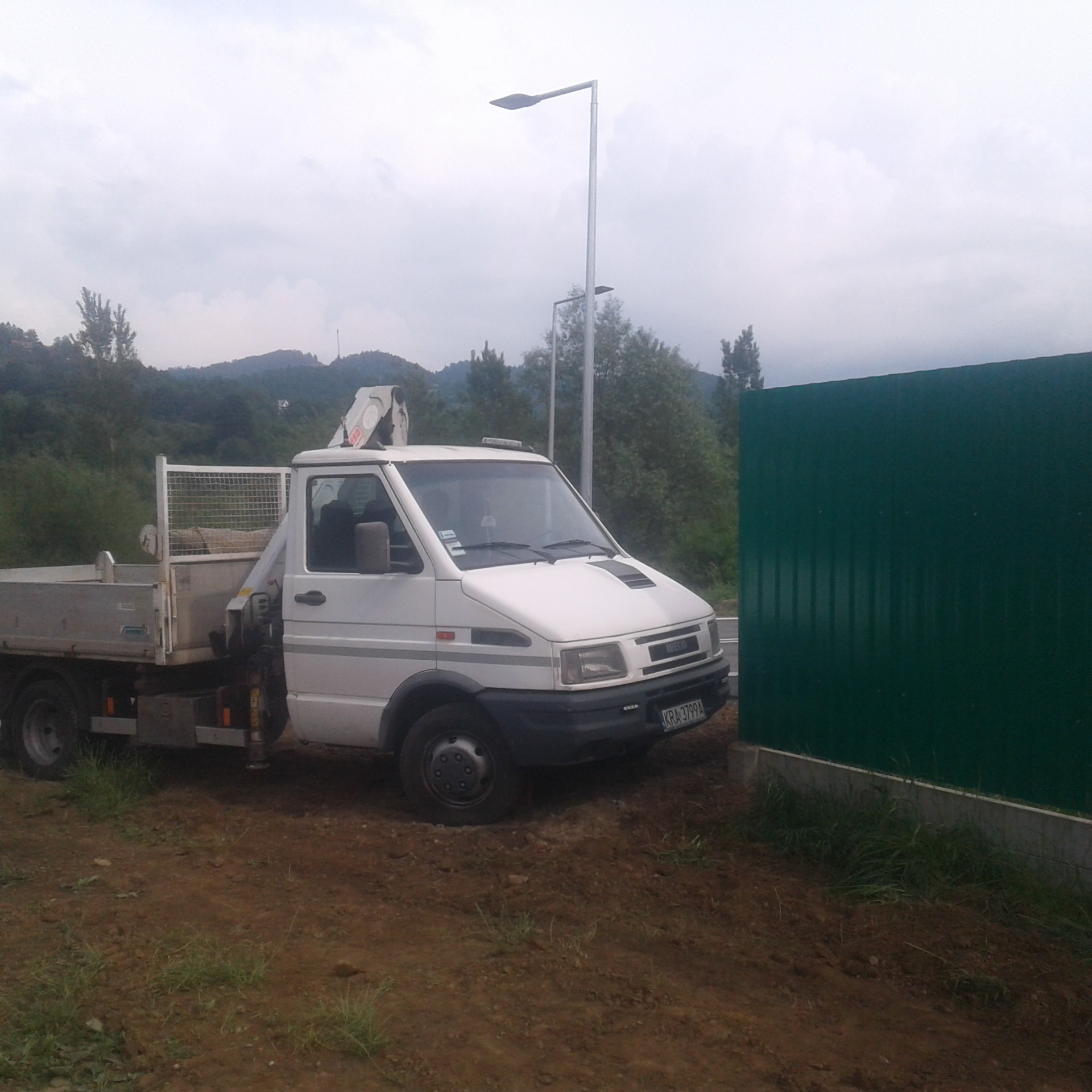 Biała ciężarówka z dźwigiem stoi na nierównym, błotnistym terenie obok zielonego ogrodzenia, w tle słupy oświetleniowe i zalesione wzgórza.