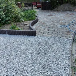 Dwa rodzaje nawierzchni: szara kostka brukowa o nieregularnym kształcie i jasny żwir oddzielone obrzeżem z tworzywa sztucznego, widoczne narzędzia ogrodnicze i wąż.