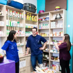 Personel w niebieskich uniformach w gabinecie z półkami pełnymi produktów dla zwierząt, widoczne karmy i akcesoria, na pierwszym planie pojemnik z przysmakami dla psów.