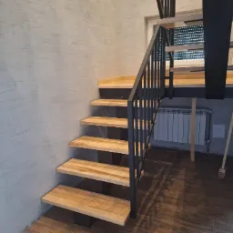 Nowoczesne schody z jasnego drewna z czarną metalową balustradą, stopnie wsparte na czarnych elementach, widoczny grzejnik za schodami, ściana o fakturze betonu i podłoga imitująca drewno.