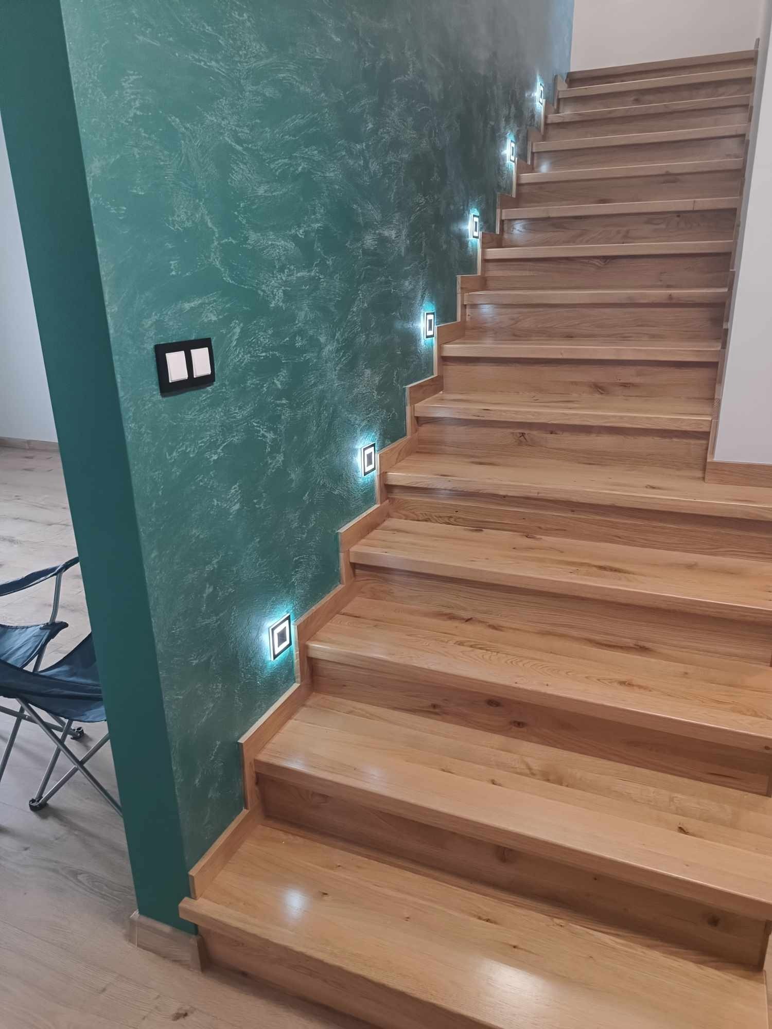 Jasne drewniane schody z podświetleniem LED w ścianie o zielonej, teksturowanej powierzchni, obok widoczny fragment krzesła turystycznego.