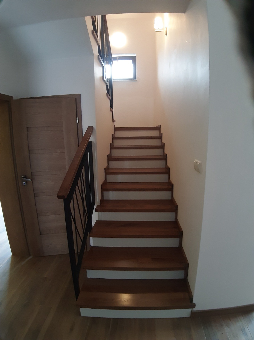 Wewnętrzne schody z drewnianymi stopniami i czarną metalową balustradą, prowadzące do góry, z widocznym oknem w tle i drzwiami po lewej stronie.