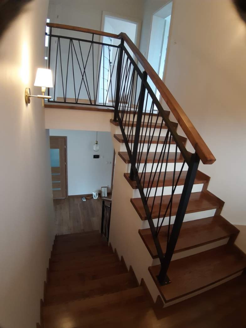 Nowoczesne schody z drewnianymi stopniami i czarną, metalową balustradą z drewnianym poręczą, oświetlone kinkietem na ścianie.