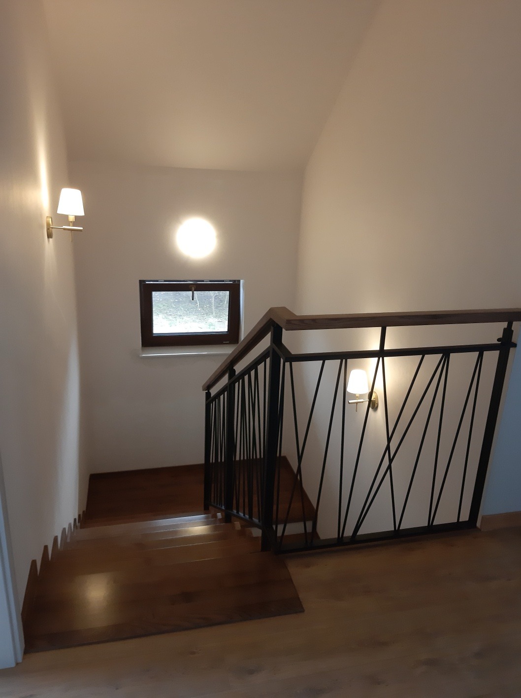 Wewnętrzne schody z drewnianymi stopniami i czarną, metalową balustradą o minimalistycznym wzorze, oświetlone kinkietami ściennymi i światłem z okna.