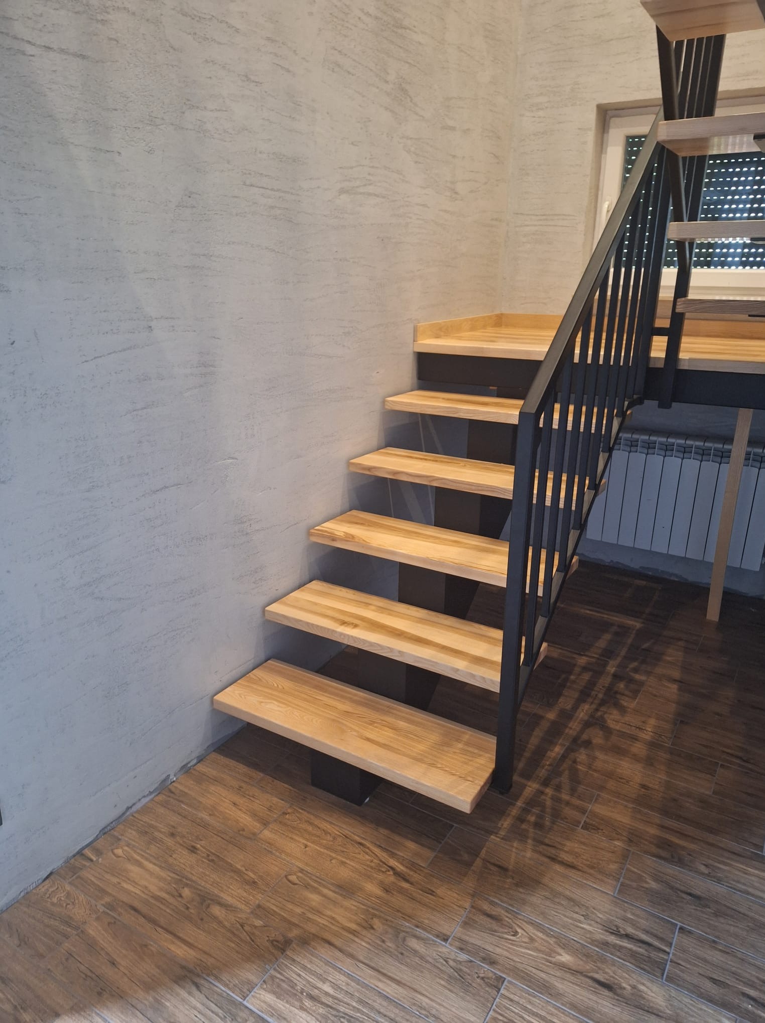 Drewniane schody z czarną metalową balustradą, zamontowane przy szarej ścianie o fakturze betonu, z widoczną podłogą z płytek imitujących drewno.