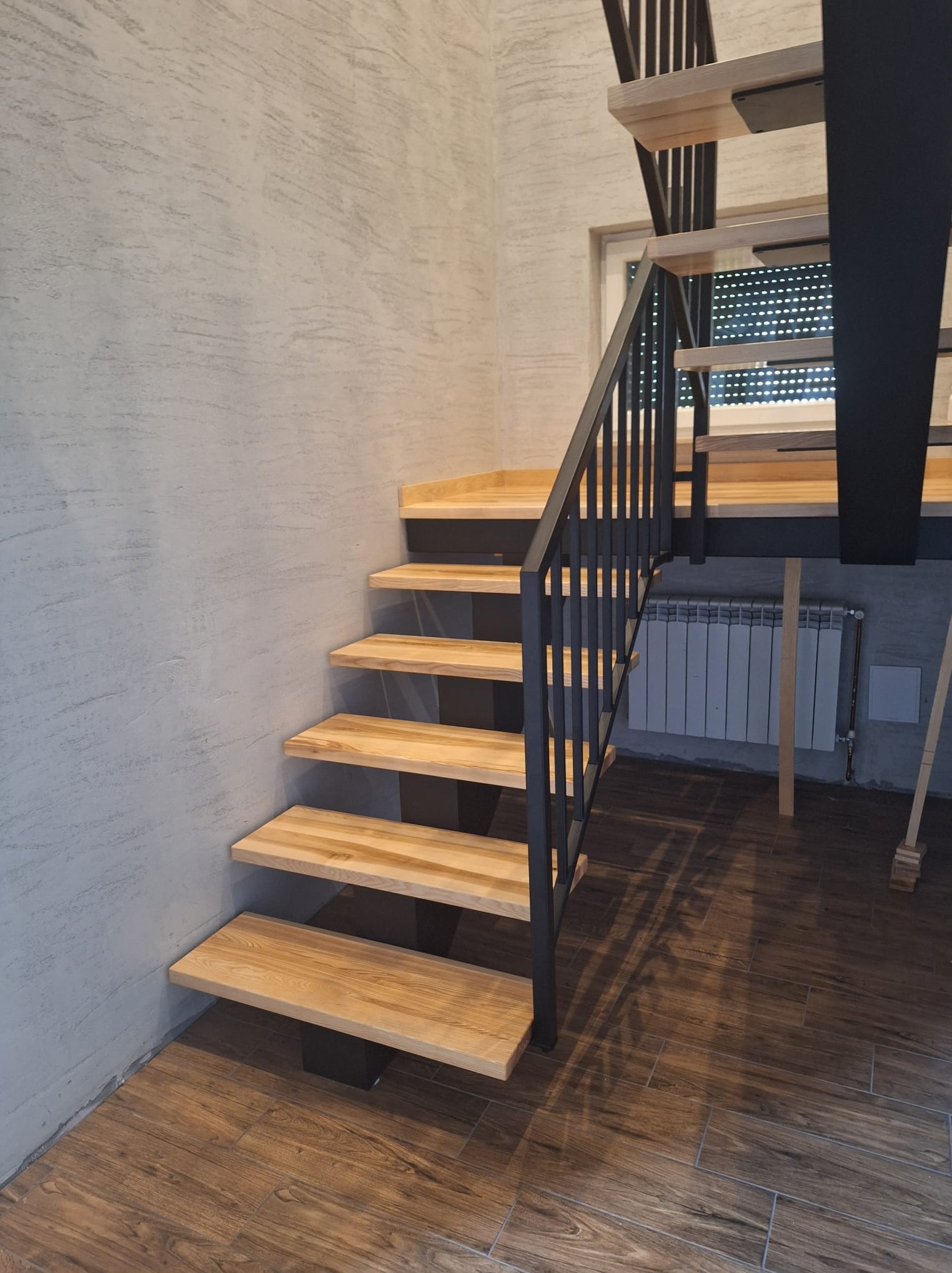 Nowoczesne schody z jasnego drewna z czarną metalową balustradą, stopnie wsparte na czarnych elementach, widoczny grzejnik za schodami, ściana o fakturze betonu i podłoga imitująca drewno.