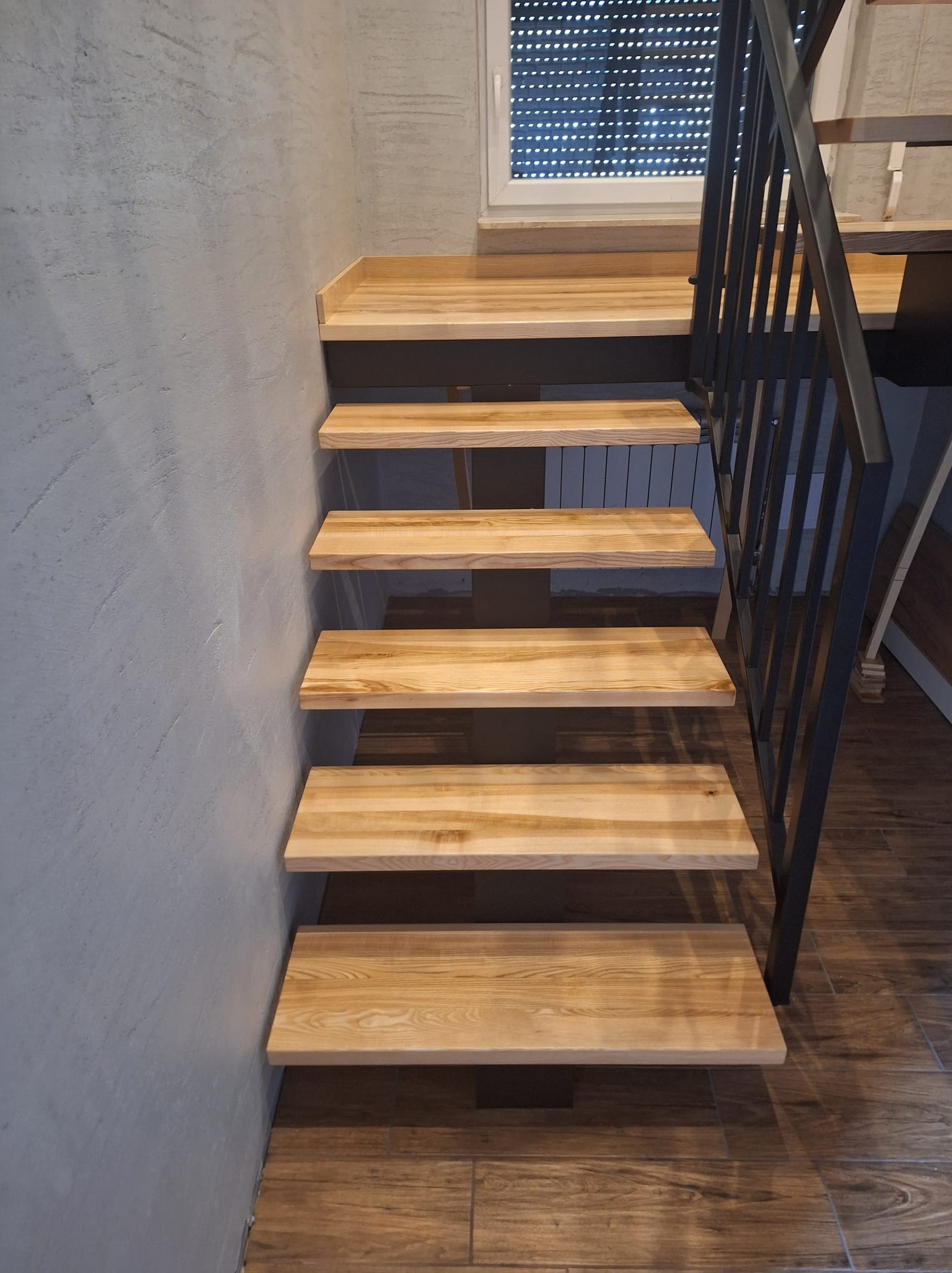 Klatka schodowa z jasnego drewna z czarną metalową balustradą, widok z dołu, ściana o szarej, chropowatej fakturze, brązowe płytki imitujące drewno na podłodze.