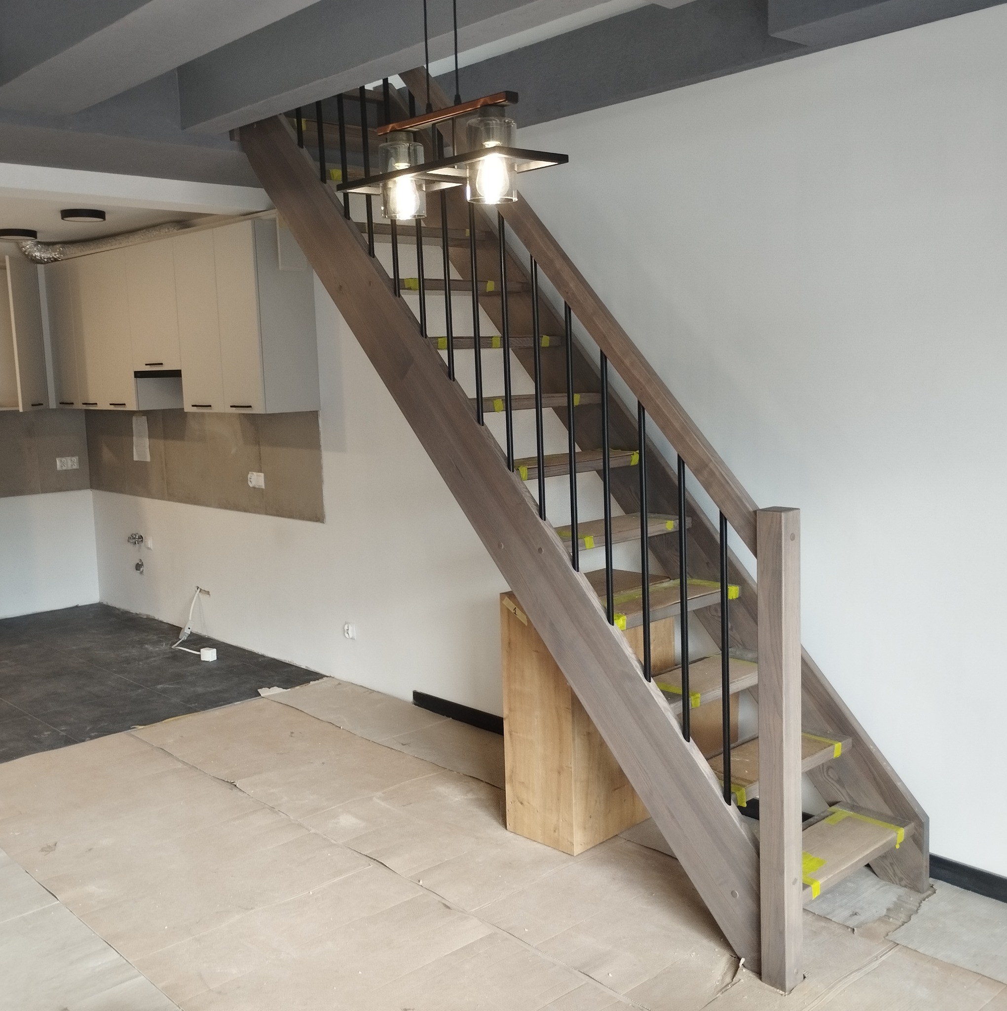 Wnętrze w trakcie remontu z zamontowanymi drewnianymi schodami z czarnymi balustradami, zabezpieczonymi żółtą taśmą, oraz lampą wiszącą.
