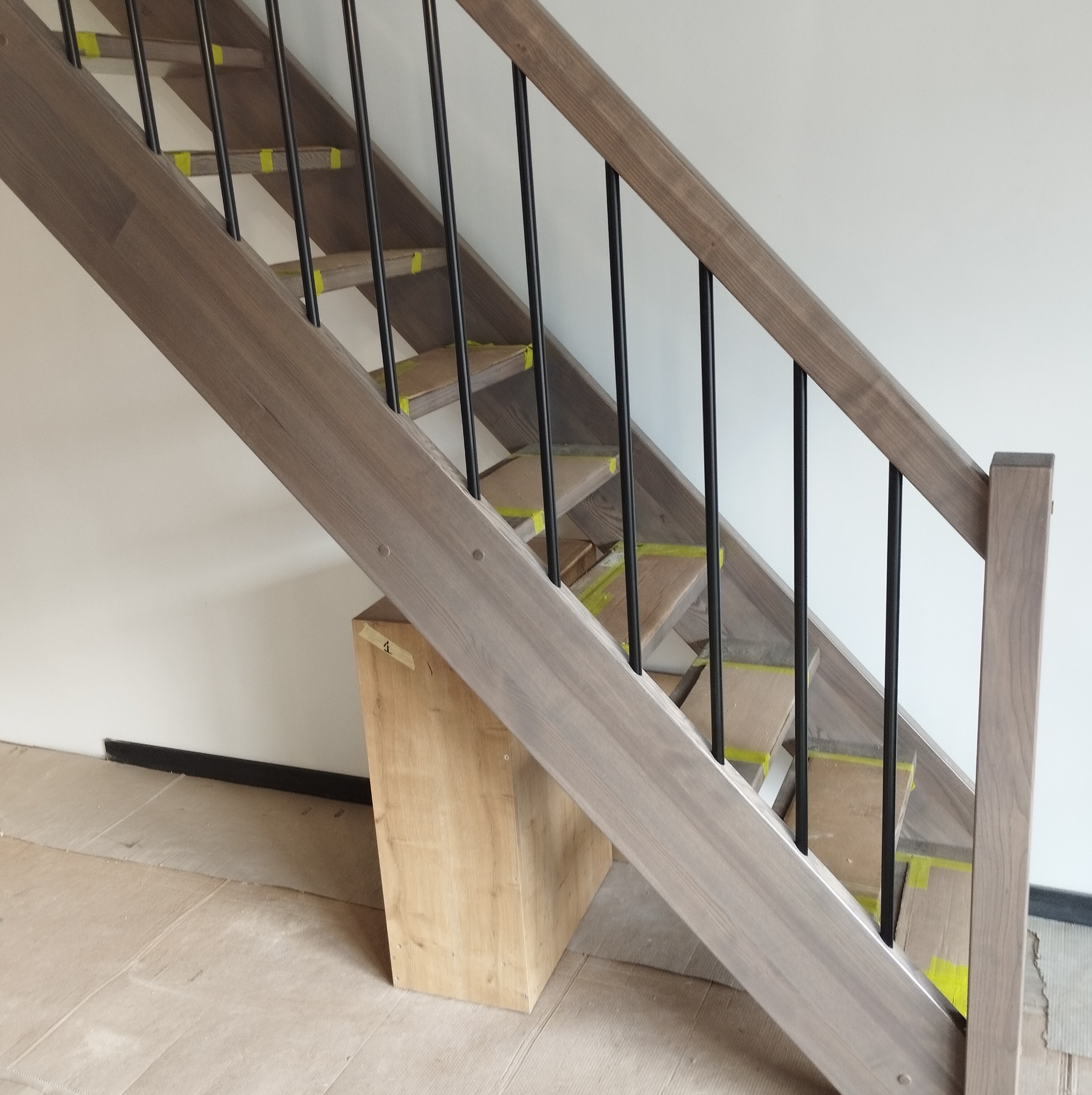 Wewnętrzne schody z drewnianymi stopniami zabezpieczonymi taśmą, czarne metalowe balustrady, widok z dołu w trakcie montażu.