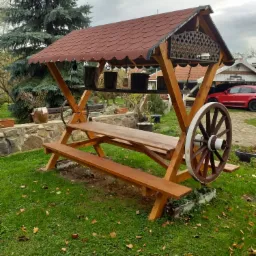 Drewniana konstrukcja z dachem krytym gontem, imitująca wóz, z kołami po bokach, służąca jako miejsce do siedzenia i spożywania posiłków na zewnątrz, ustawiona w ogrodzie.