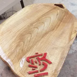 Drewniana taca z rzeźbionym, czerwonym, orientalnym znakiem, widoczna na stole warsztatowym z narzędziami w tle.