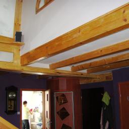 DOCTORWOOD Leszek Lis https://doctorwoodsklep.pl/ - Wnętrze domu z drewnianą konstrukcją stropu, widoczne belki, schody z balustradą z surowego drewna oraz elementy dekoracyjne jak zegar i regał na ścianie w kolorze fioletowym.
