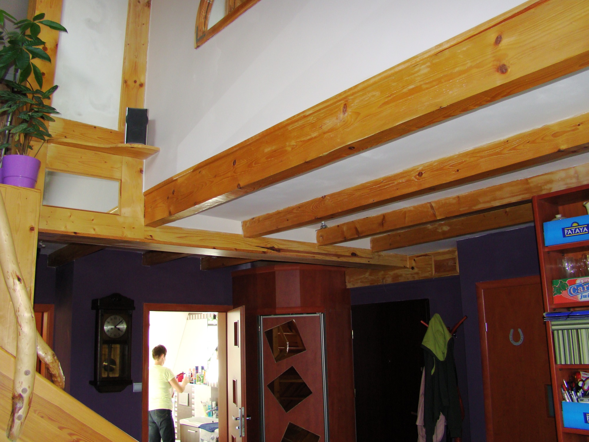 Wnętrze domu z drewnianą konstrukcją stropu, widoczne belki, schody z balustradą z surowego drewna oraz elementy dekoracyjne jak zegar i regał na ścianie w kolorze fioletowym.