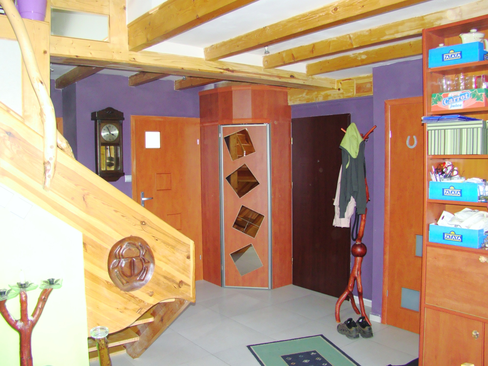 Wnętrze domu z drewnianymi schodami z dekoracyjnym płaskorzeźbą, drewnianymi belkami stropowymi, lustrzanymi drzwiami i drewnianym wieszakiem na ubrania w kształcie drzewa.