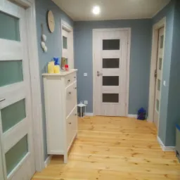 Przedpokój z drewnianą podłogą, trzema parami drzwi z przeszkleniami, komodą na buty, zegarem i dekoracjami na ścianie w kolorze gołębiej szarości.