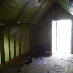 Poddasze w trakcie ocieplania, widoczne folie paroizolacyjne na ścianach i suficie, narzędzia na podłodze, otwarte drzwi z jasnym światłem wpadającym do środka.