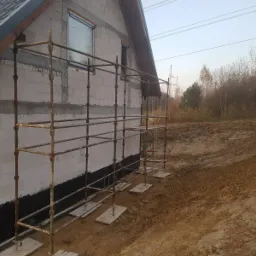 Dom w trakcie ocieplania, widoczne rusztowania, bloczki z betonu komórkowego, izolacja przeciwwilgociowa przy gruncie oraz okna z ramami w kolorze antracytowym.