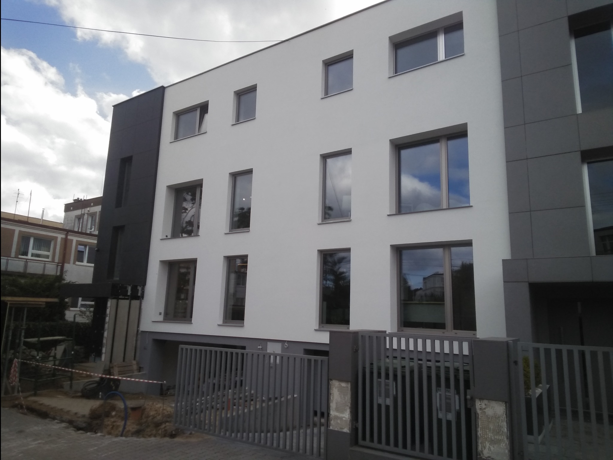 Biało-szara elewacja budynku wielorodzinnego z widocznymi oknami i szarą bramą wjazdową, teren wokół budynku w trakcie prac wykończeniowych.