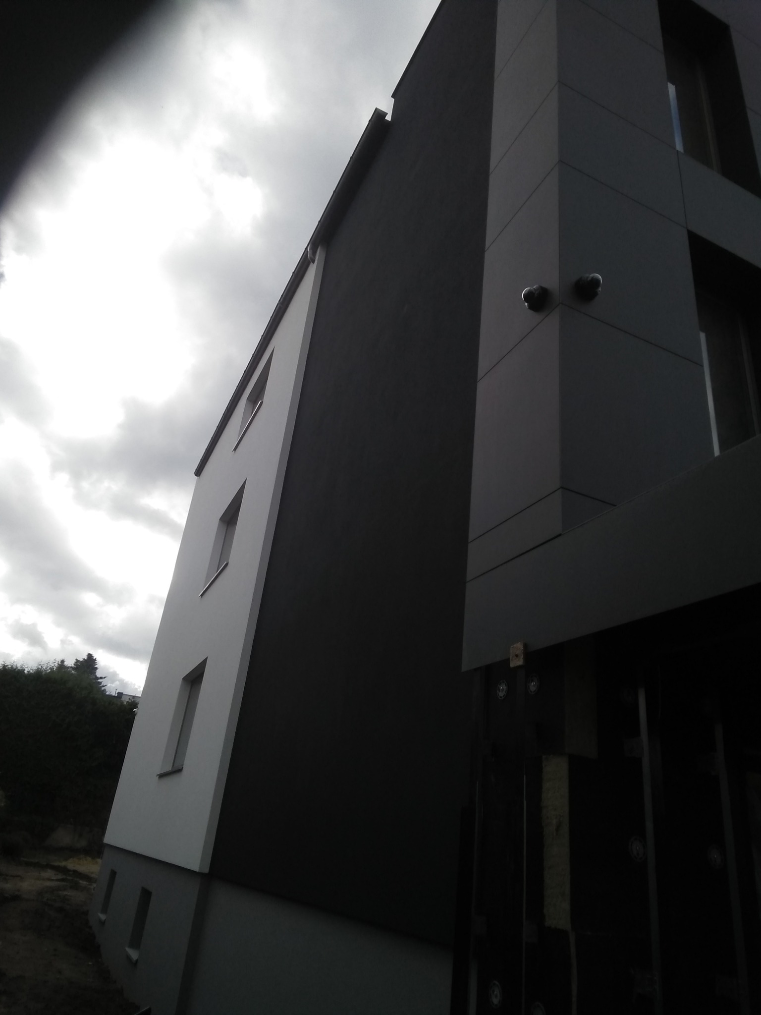 Nowoczesny budynek z elewacją w odcieniach szarości i bieli, widok z dołu na pochmurne niebo, z widocznymi elementami montażowymi i kamerami.