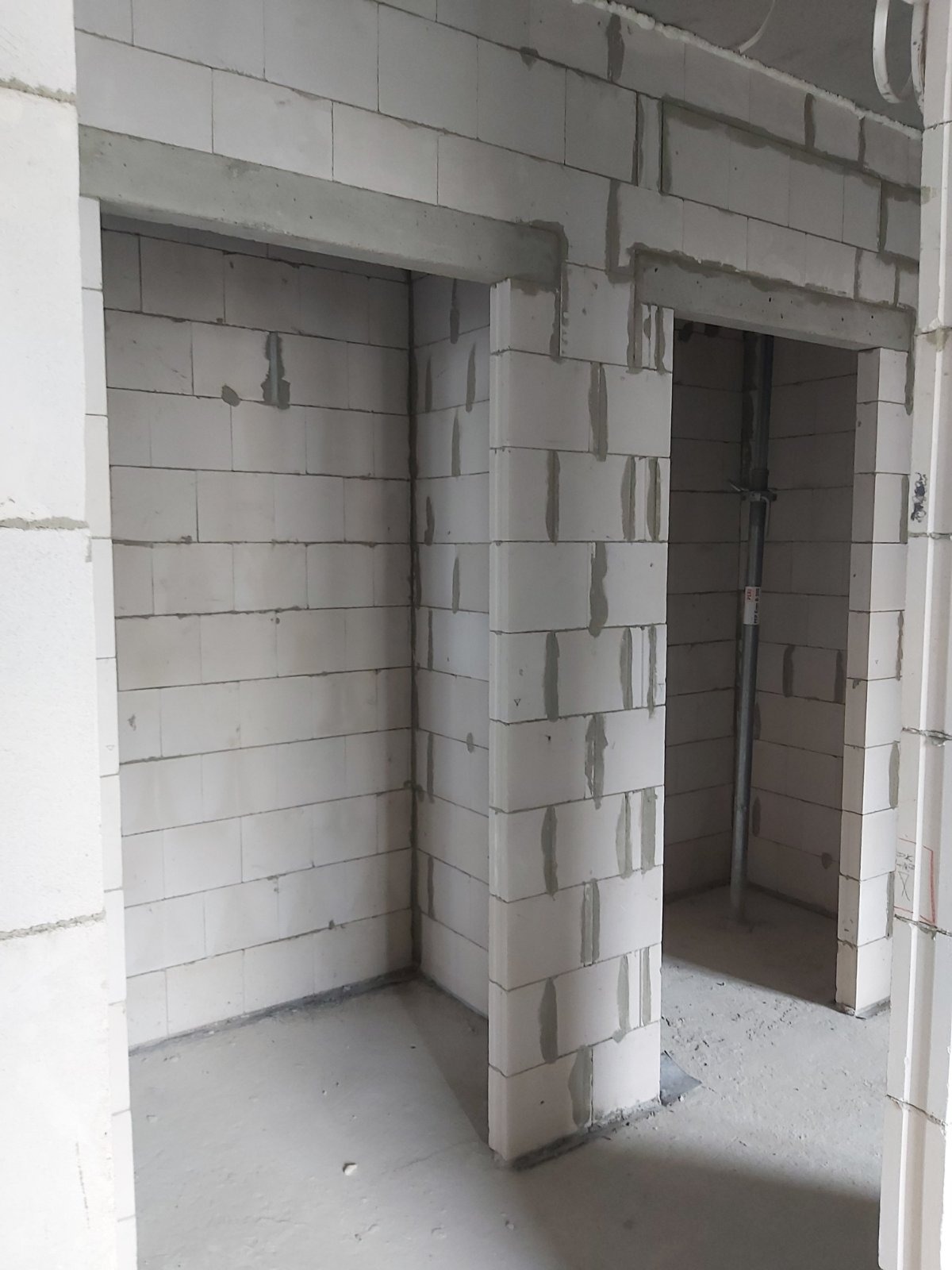 Surowe wnętrze w trakcie budowy z białych bloczków, widoczne dwa otwory drzwiowe z betonowymi nadprożami i metalowa rura podtrzymująca strop.