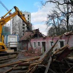 Żółta koparka JCB z łyżką na wysięgniku demontuje fragment dachu ceglanego, częściowo zburzonego budynku. W tle widać nowe budynki w trakcie budowy i nagie drzewa.