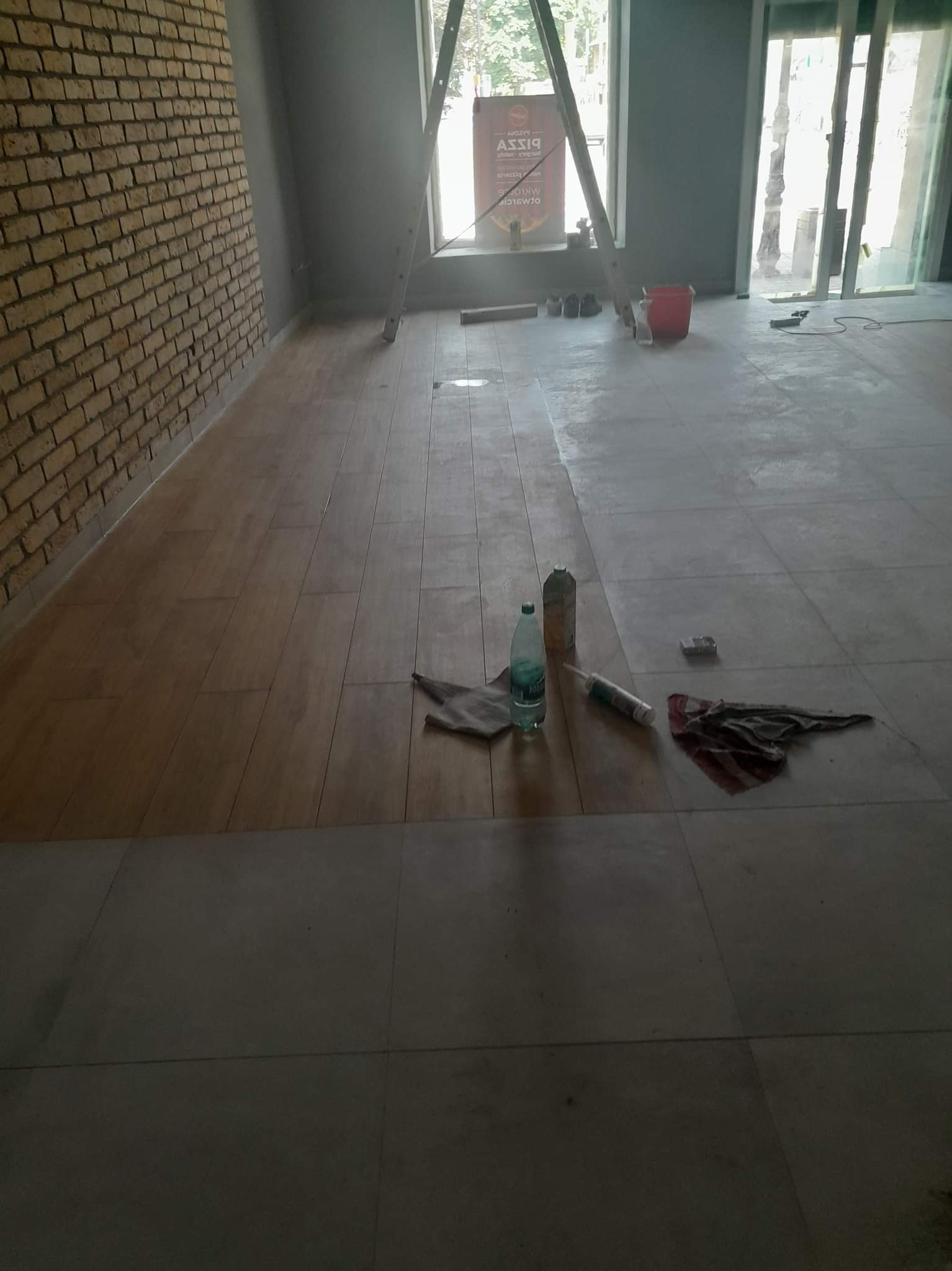 Wnętrze w trakcie prac wykończeniowych: częściowo ułożona podłoga z płytek imitujących drewno, widoczna ceglana ściana, drabina oparta o okno, butelka wody, silikon i szmatka na podłodze.