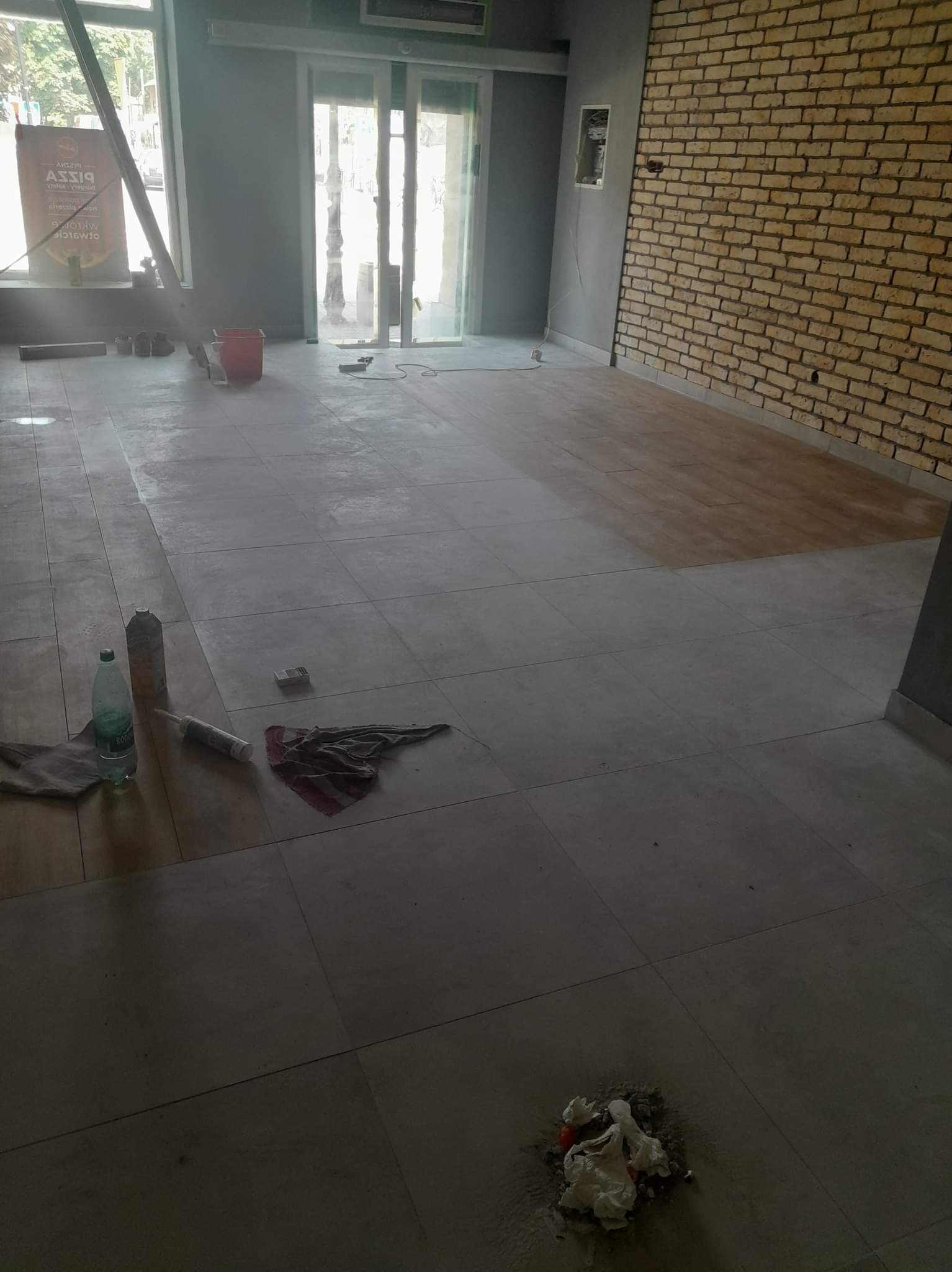 Wnętrze lokalu w trakcie prac wykończeniowych: częściowo ułożona podłoga z płytek, fragment ściany z cegły, butelka wody, tuba z klejem, szmata i resztki zaprawy na podłodze.