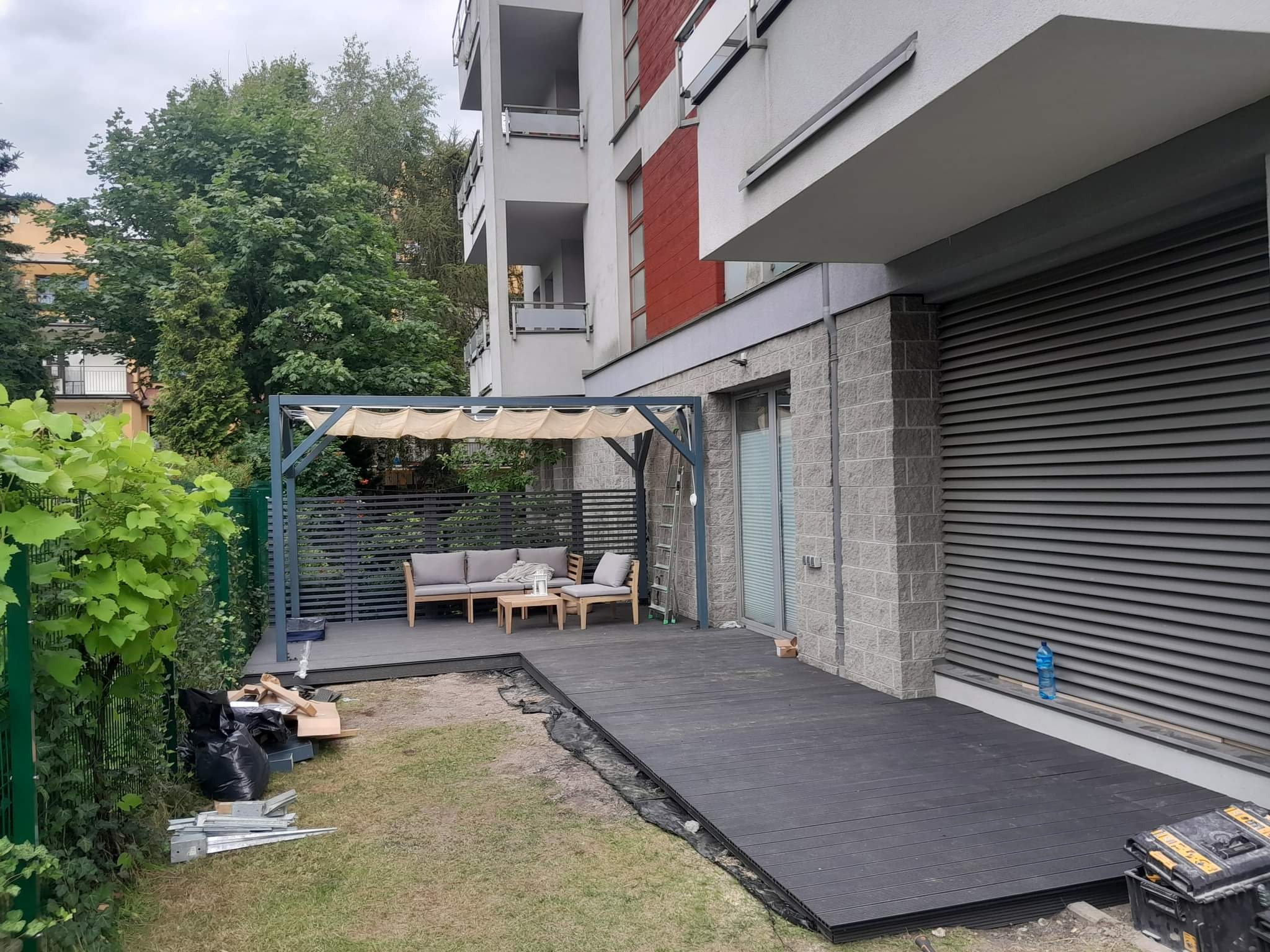 Montaż tarasu kompozytowego przy nowoczesnym budynku, widoczne deski, narzędzia i elementy konstrukcyjne, częściowo zagospodarowana przestrzeń z meblami ogrodowymi i pergolą, teren wokół w trakcie...