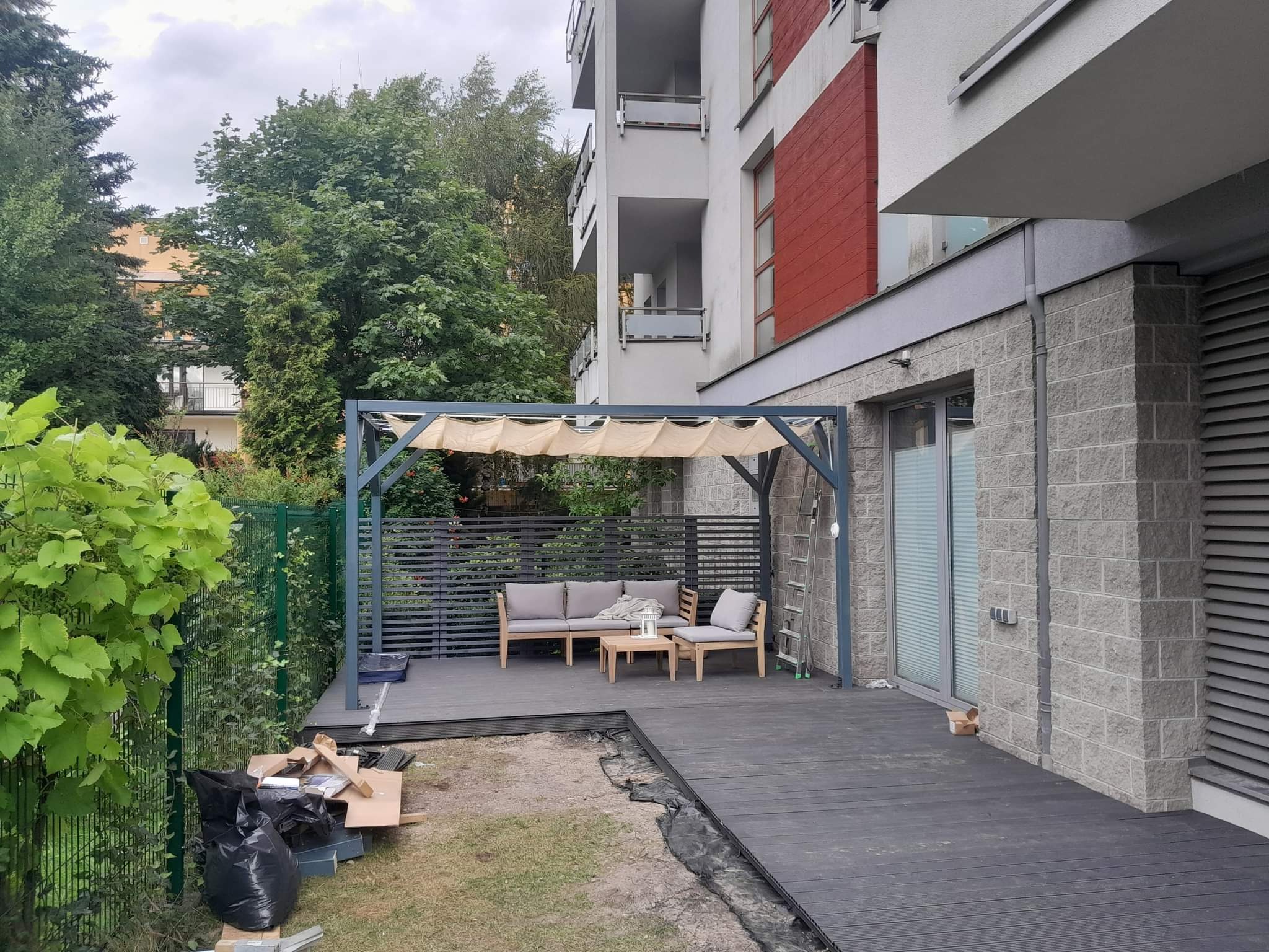 Częściowo wykończony taras z kompozytowych desek, szara pergola z beżowym zadaszeniem, meble ogrodowe, widoczne ślady prac budowlanych, budynek mieszkalny w tle.