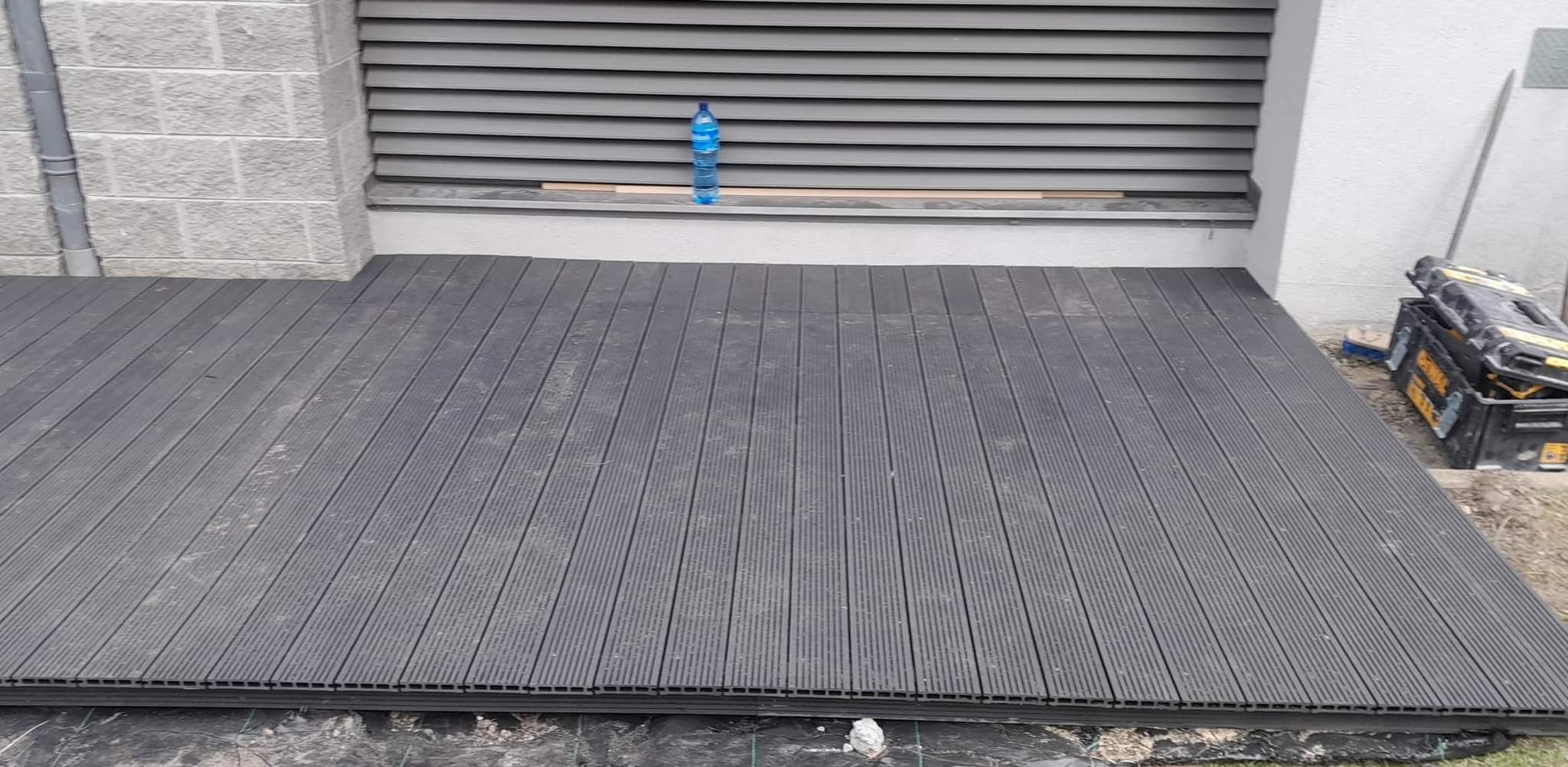 Szara deska tarasowa ułożona na folii, z butelką wody i skrzynkami narzędziowymi w tle.